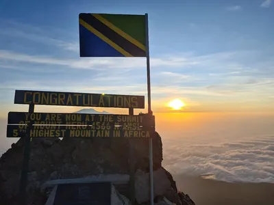 Congratulations summit sign at Mount Meru's peak, 4,566m, with Tanzanian flag and sunrise over the clouds